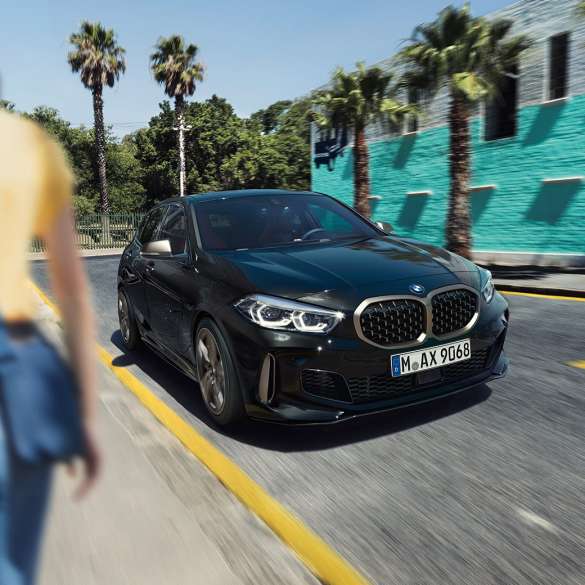 BMW M135i xDrive, háromnegyedes elölnézet. Egy Zafírfekete metál BMW M135i xDrive háromnegyedes elölnézetből, egy nővel az előtérben.
