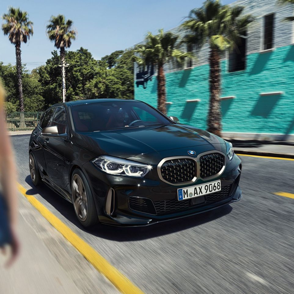 BMW M135i xDrive, háromnegyedes elölnézet. Egy mozgó, Zafírfekete metálfényezésű BMW M135i xDrive háromnegyedes elölnézetből, egy nővel az előtérben.