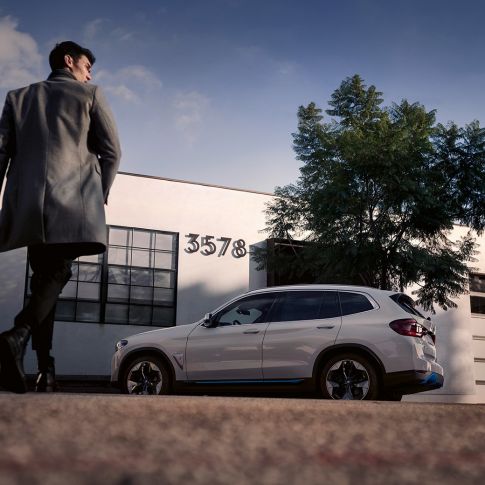 BMW iX3, G08, oldalnézet egy épület előtt, egy férfival BMW iX3, G08, 2020, egy épület előtt álló, Kristály fehér metálfényezésű autó oldalnézetből, egy férfival