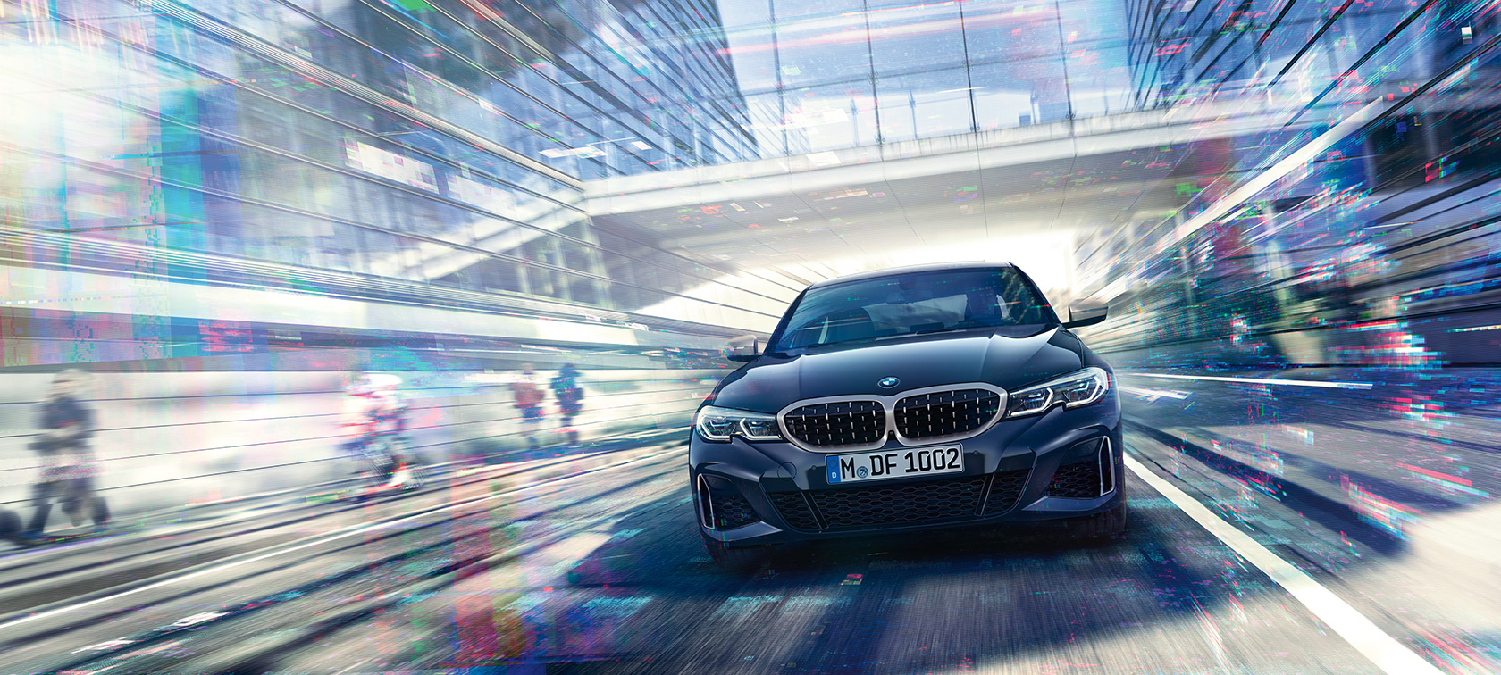 BMW 3-as Limousine, egy BMW M340i xDrive elölnézetből. Egy BMW M340i xDrive Limousine üvegborítású városi épületek között halad.