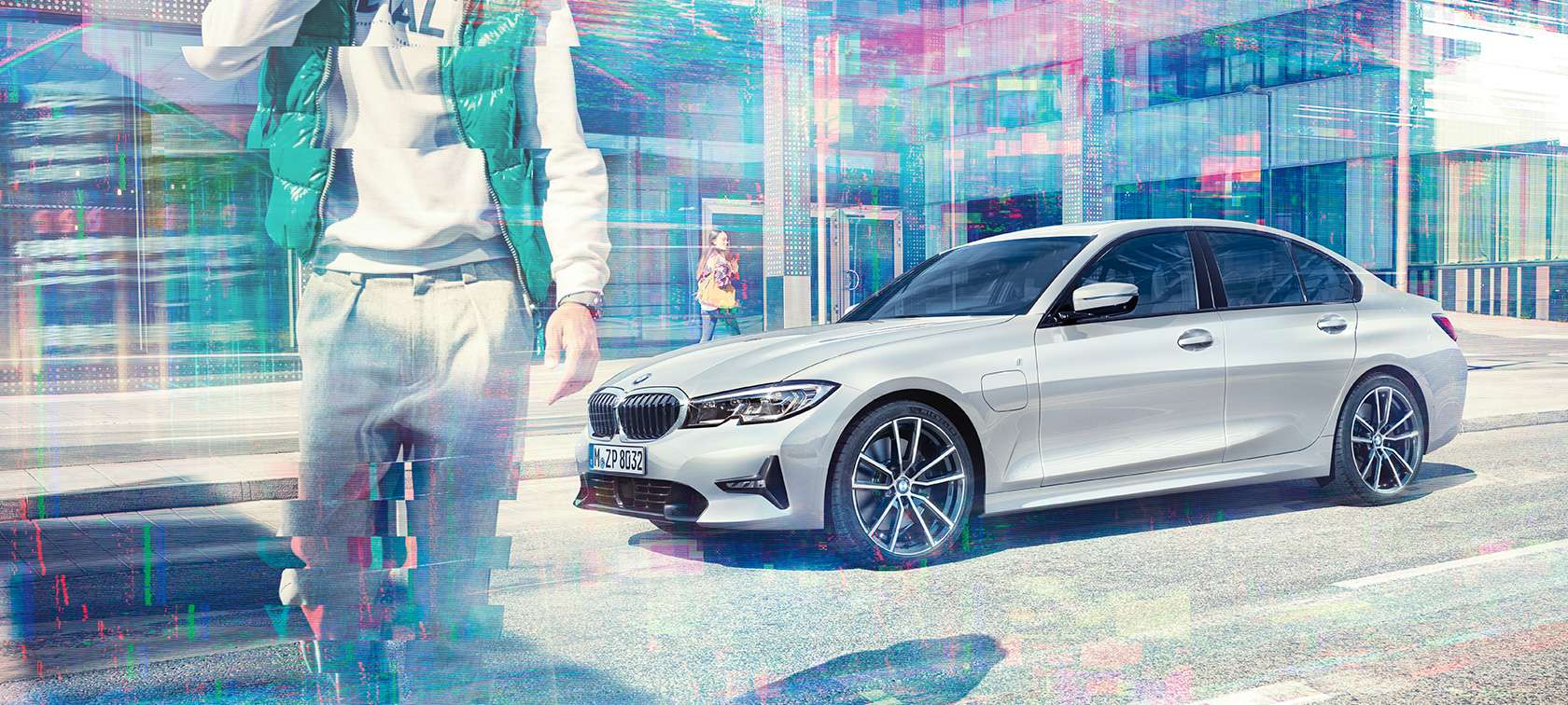 BMW 3-as Limousine, a BMW 330e oldalnézetből. Egy álló BMW 330e Limousine háromnegyedes oldalnézetből, egy üvegborítású városi épület előtt, sétáló utasokkal.