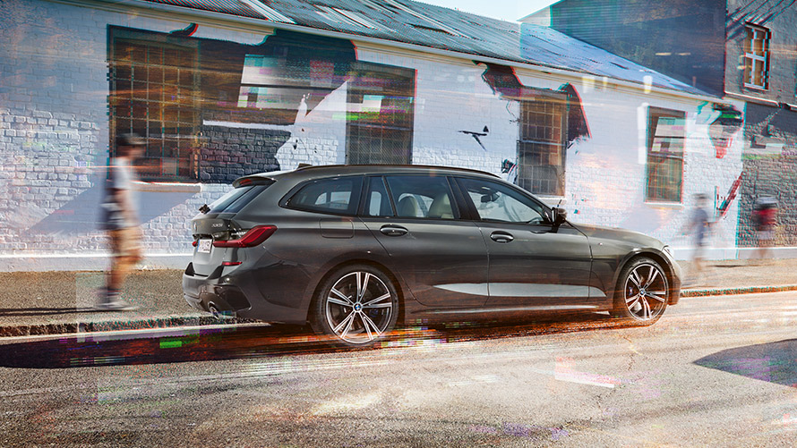 BMW 3 Touring (G21), Dravit szürke, hátulnézet BMW 3 Touring (G21), egy épület előtt elhaladó, Dravit szürke autó rézsútos hátulnézetből