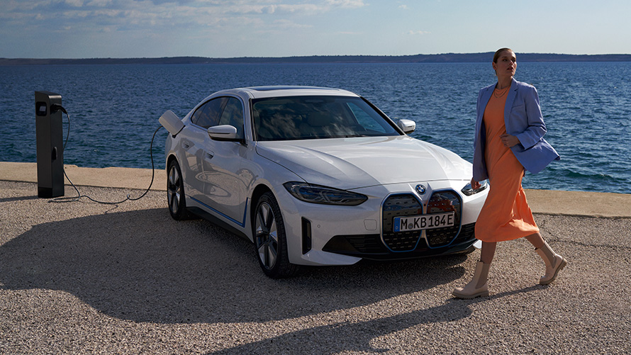 Rézsútos elölnézet, BMW i4 eDrive40, G26, 2021, egy töltőállomáson tartózkodó autó Egy nő áll egy Kristály fehér, rézsútos elölnézetből látható, töltőállomáson tartózkodó BMW i4 eDrive40 (G26) előtt, tóval a háttérben