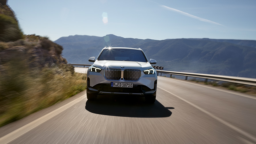 BMW IX1, U11 BEV, elölnézet BMW iX1, U11 BEV, egy hegyi úton haladó, Kristályfehér metálfényezésű autó