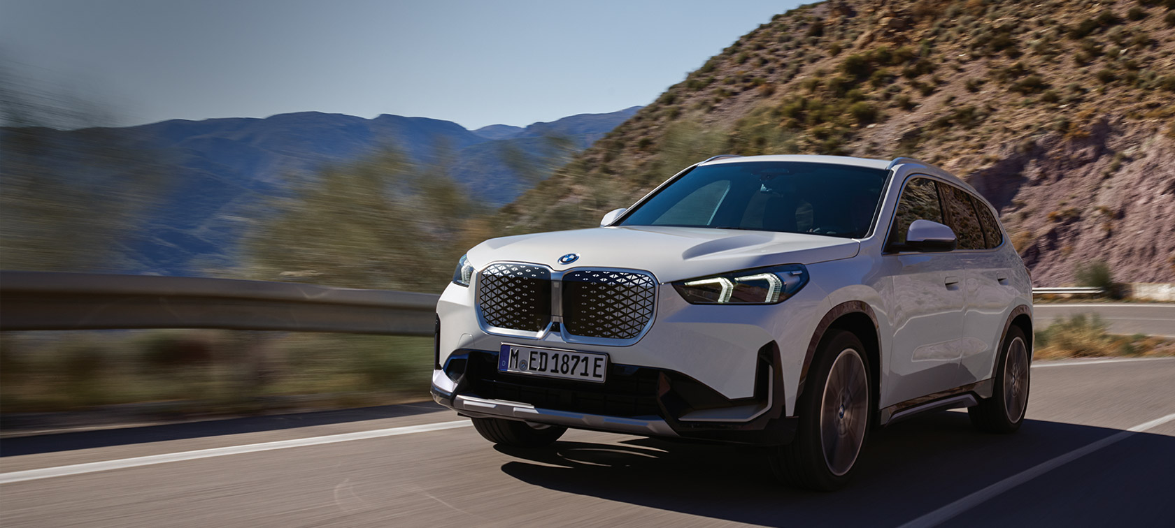 BMW iX1, U11 BEV, egy hegyi úton haladó, Kristályfehér metálfényezésű autó rézsútos elölnézetből