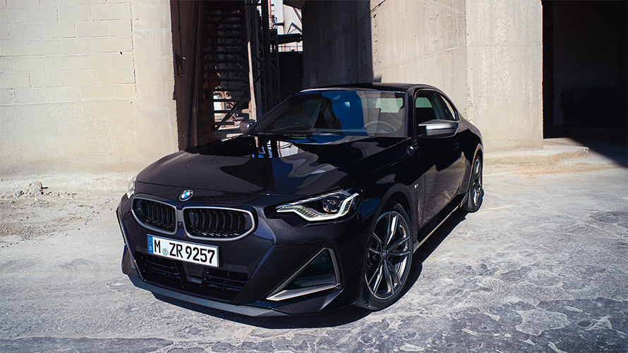 BMW M240i xDrive Coupé, G42, 2021, egy álló autó rézsútos elölnézetből BMW M240i xDrive Coupé, G42, 2021, egy raktár előtt álló, Zafírfekete metálfényezésű autó rézsútos elölnézetből