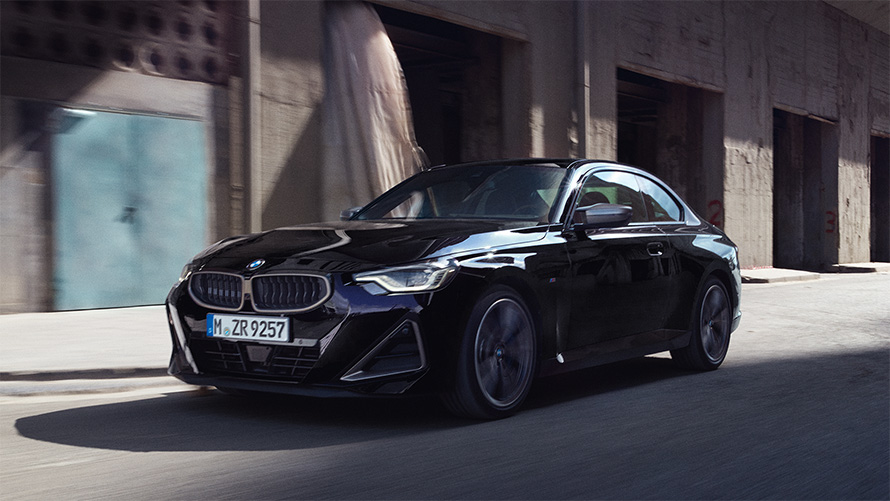 BMW M240i xDrive Coupé, G42, 2021, egy mozgó autó rézsútos oldalnézetből BMW M240i xDrive Coupé, G42, 2021, egy raktárépületek előtt elhaladó, Zafírfekete metálfényezésű autó rézsútos oldalnézetből