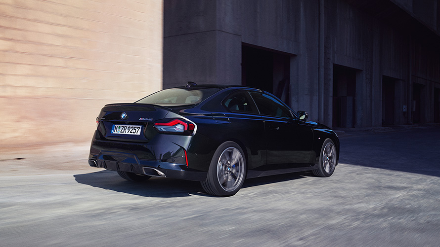BMW M240i xDrive Coupé, G42, 2021, egy álló autó rézsútos hátulnézetből BMW M240i xDrive Coupé, G42, 2021, egy raktár előtt álló, Zafírfekete metálfényezésű autó rézsútos hátulnézetből