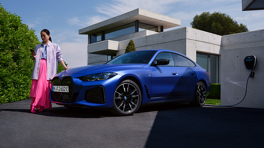BMW i4 M50, G26, 2021, egy ház előtt álló, egy nő társaságában látható BMW Individual Frozen Portimao kék metálfényezésű autó rézsútos elölnézetből