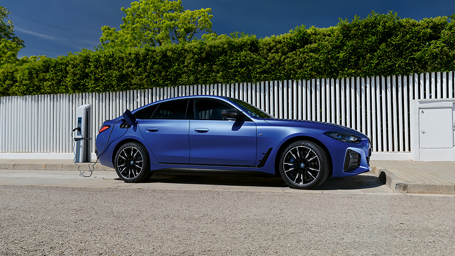 BMW i4, M50, G26, 2021, egy töltőoszlophoz csatlakoztatott BMW Individual Frozen Portimao kék metálfényezésű autó oldalnézetből