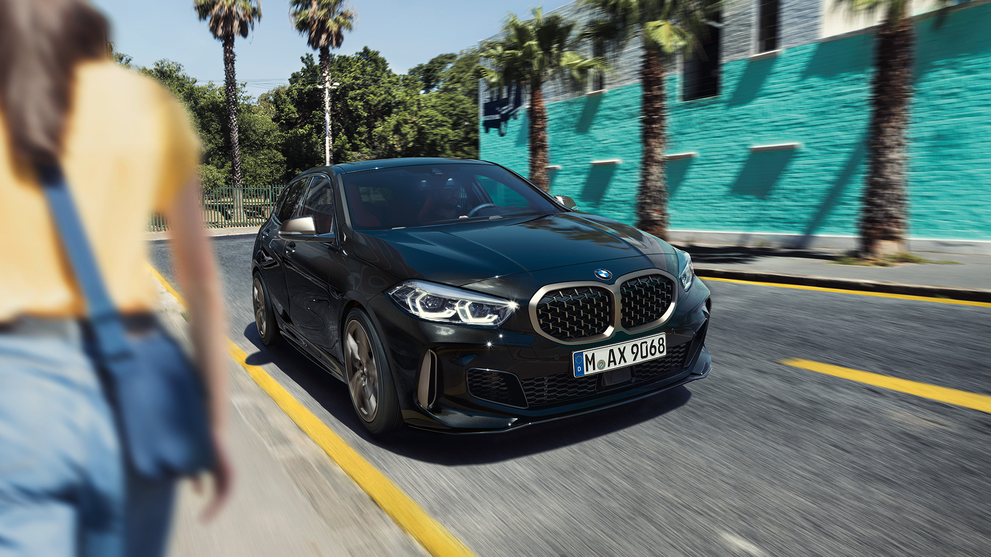 BMW M135i xDrive, háromnegyedes elölnézet. Egy Zafírfekete metál BMW M135i xDrive háromnegyedes elölnézetből, egy nővel az előtérben.