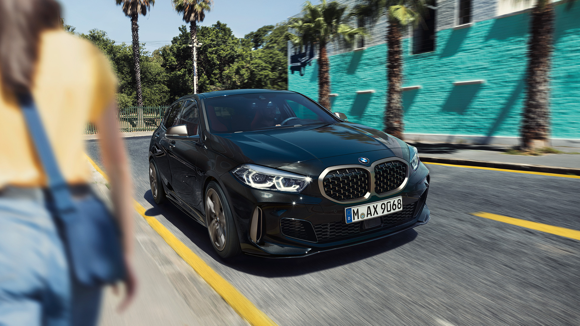 BMW M135i xDrive, háromnegyedes elölnézet. Egy mozgó, Zafírfekete metálfényezésű BMW M135i xDrive háromnegyedes elölnézetből, egy nővel az előtérben.