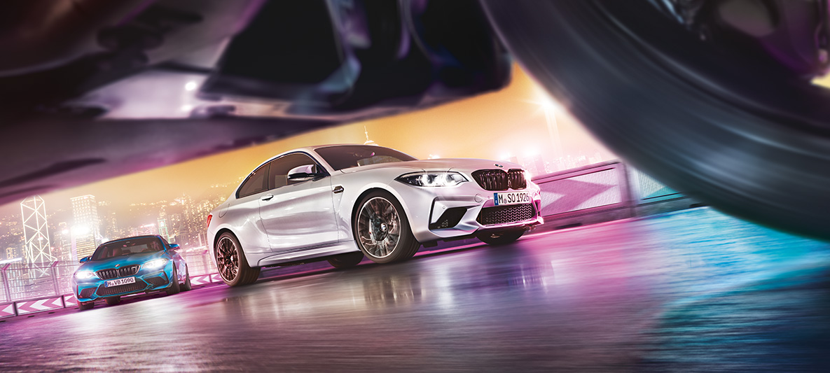 A BMW M2 Competition tükröződő aszfalton Két BMW M2 Competition a versenypályán az éjszakai várossal a háttérben, utcanézetből.