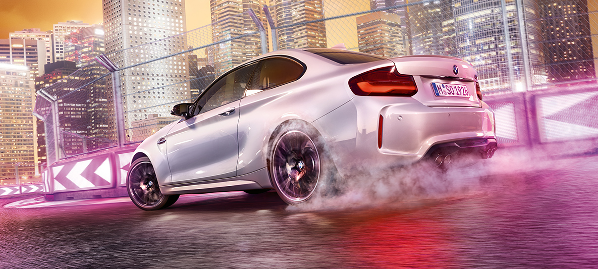 A BMW M2 Competiton gyorsít Egy fehér BMW M2 Competition driftel a versenypályán az éjszakai város sziluettjével a háttérben.