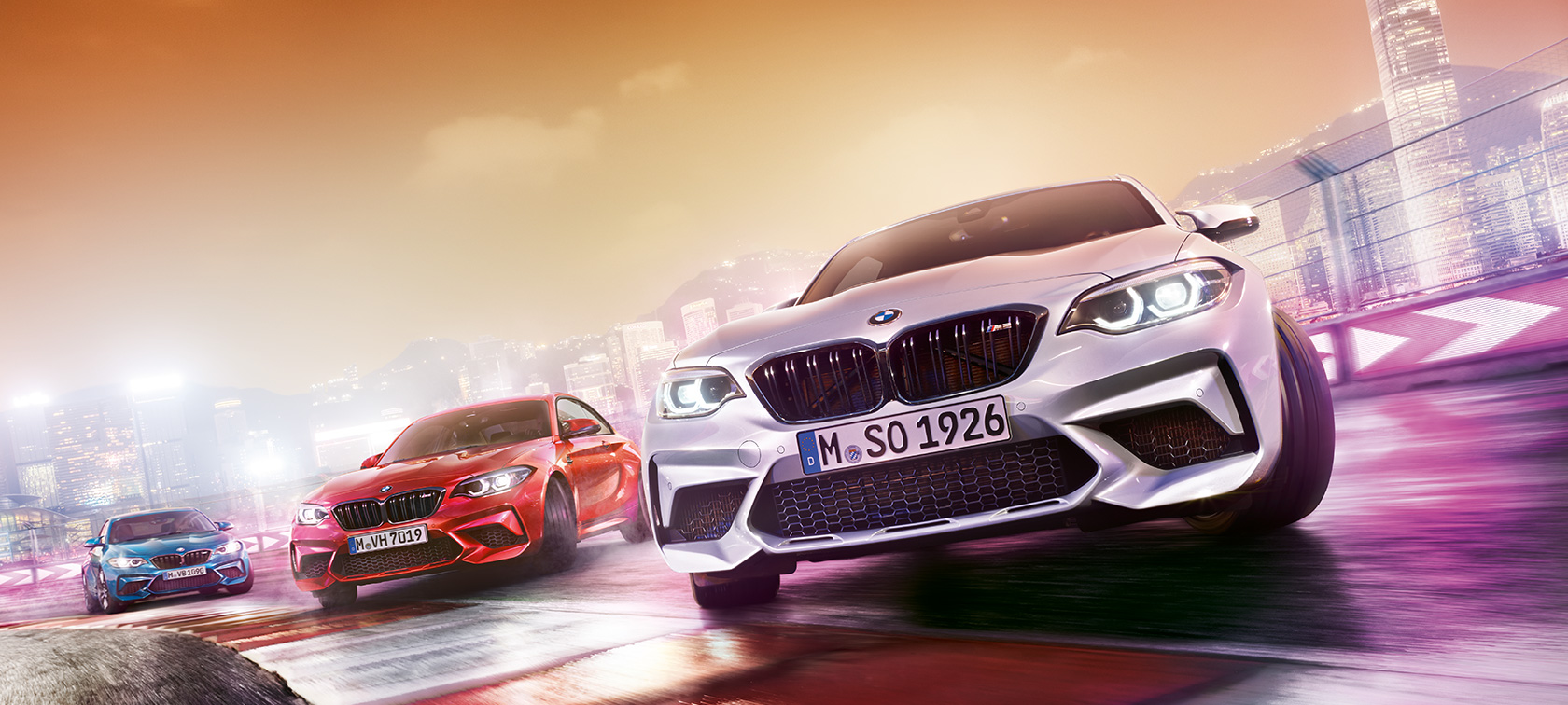 A BMW M2 Competition akció közben Három BMW M2 Competition driftel a versenypályán, a háttérben az éjszakai város.