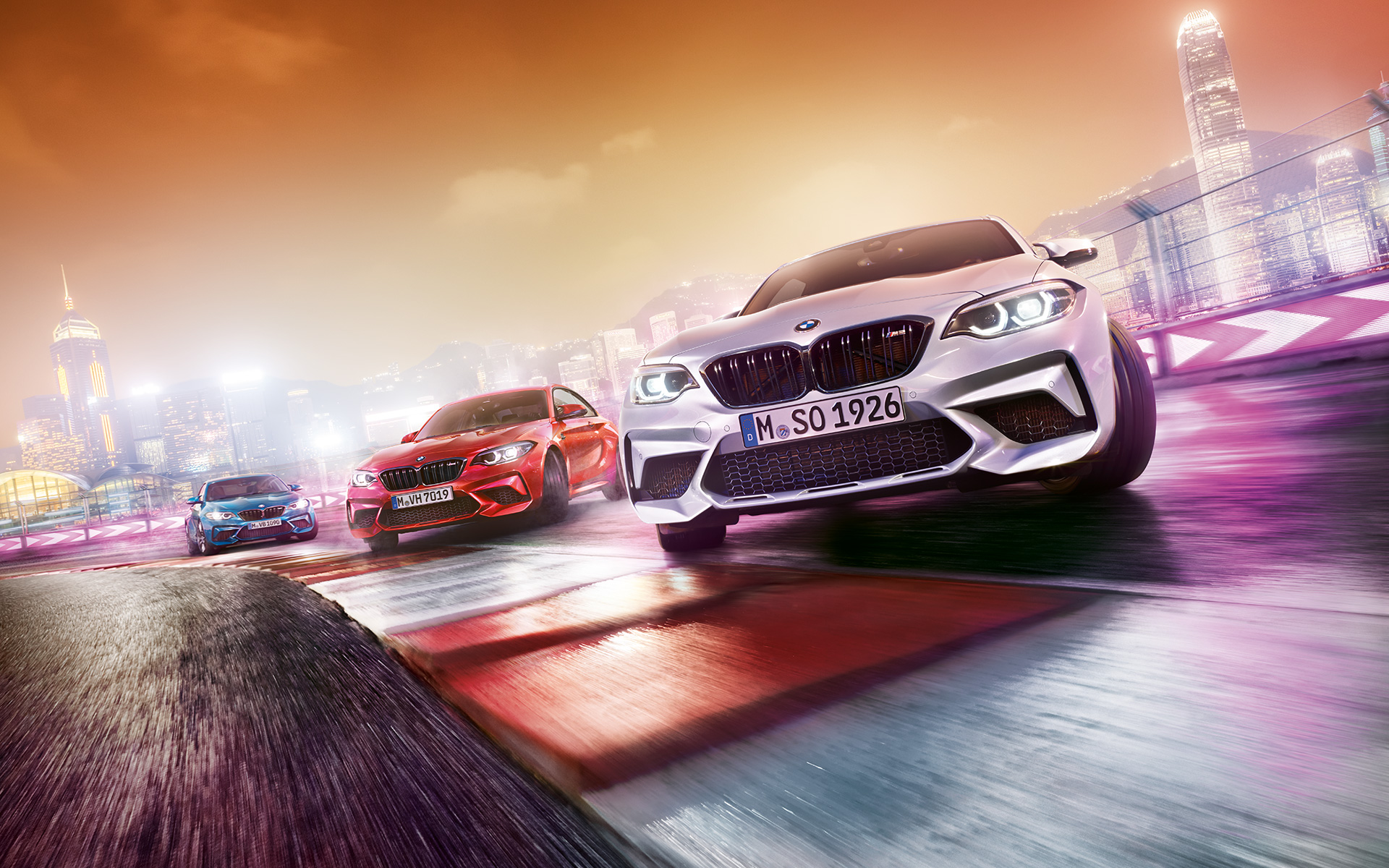 A BMW M2 Competition akció közben Három, hátulnézetből látható BMW M2 Competiton száguld a versenypályán a város éjjeli sziluettjével a háttérben.