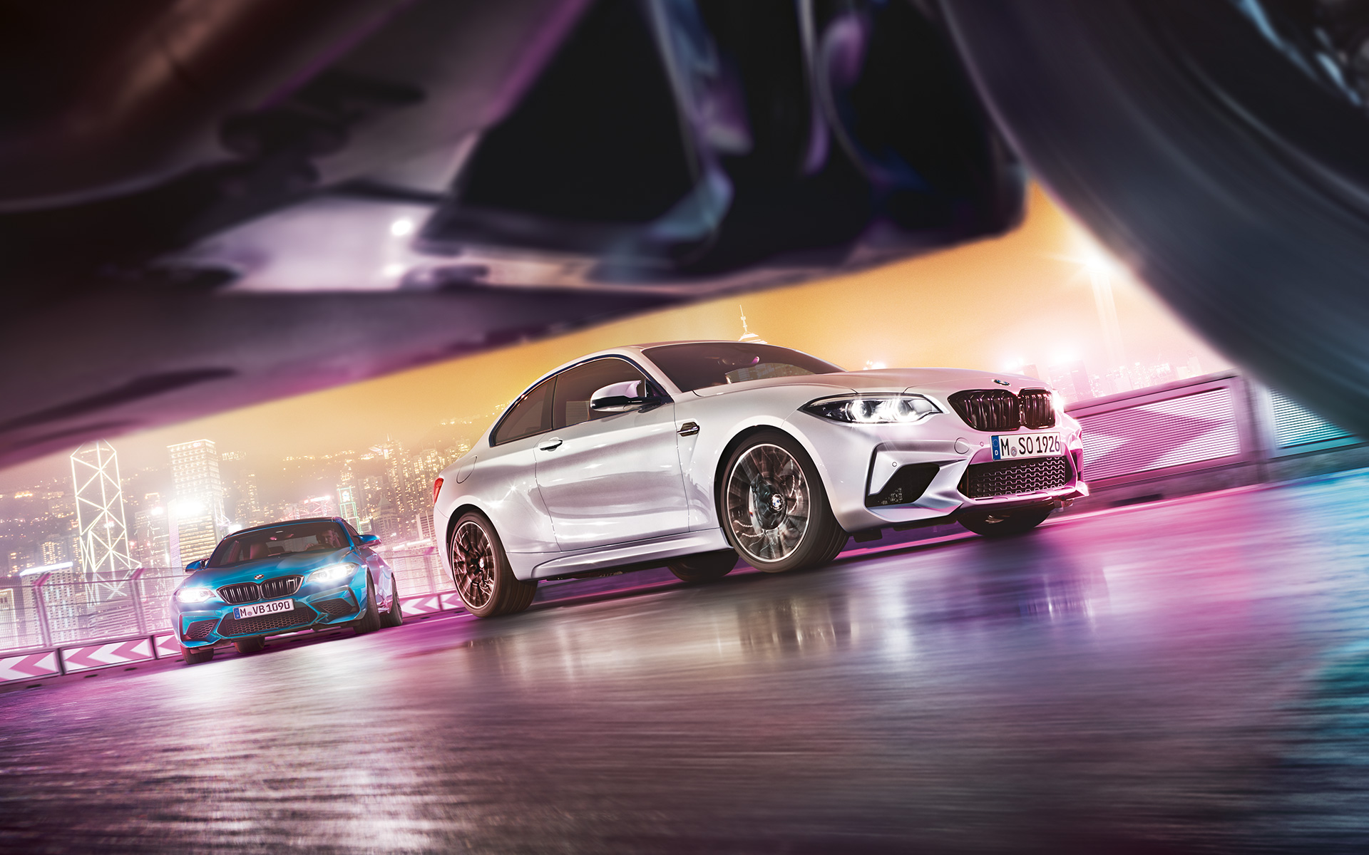 A BMW M2 Competition tükröződő aszfalton Két BMW M2 Competition a versenypályán az éjszakai várossal a háttérben, utcanézetből.