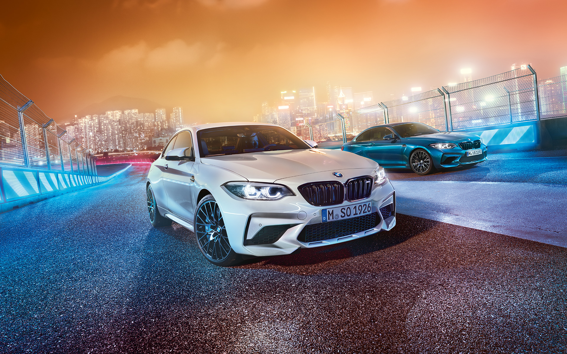 Két BMW M2 Competition a versenypályán Egy fehér és egy kék, álló BMW M2 Competition látható félprofilból, a háttérben az éjszakai város látképe.