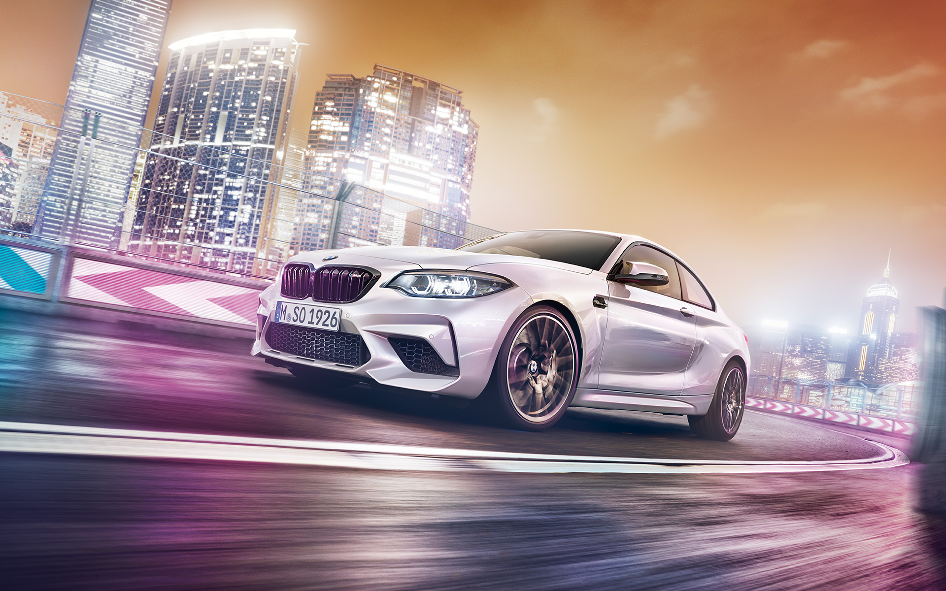 Egy fehér BMW M2 Competition a város sziluettjével Egy fehér BMW M2 Competition száguld a versenypályán az éjszakai város sziluettjével a háttérben.