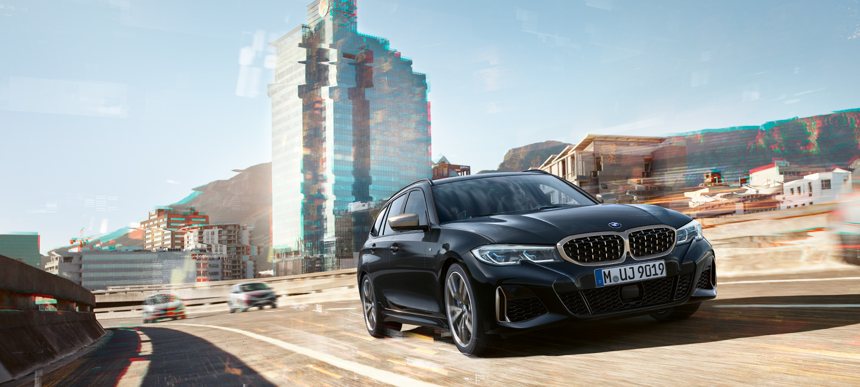 Rézsútos elölnézet városi környezetben Egy mozgó BMW M340i xDrive Touring rézsútos elölnézetből, városi környezetben.