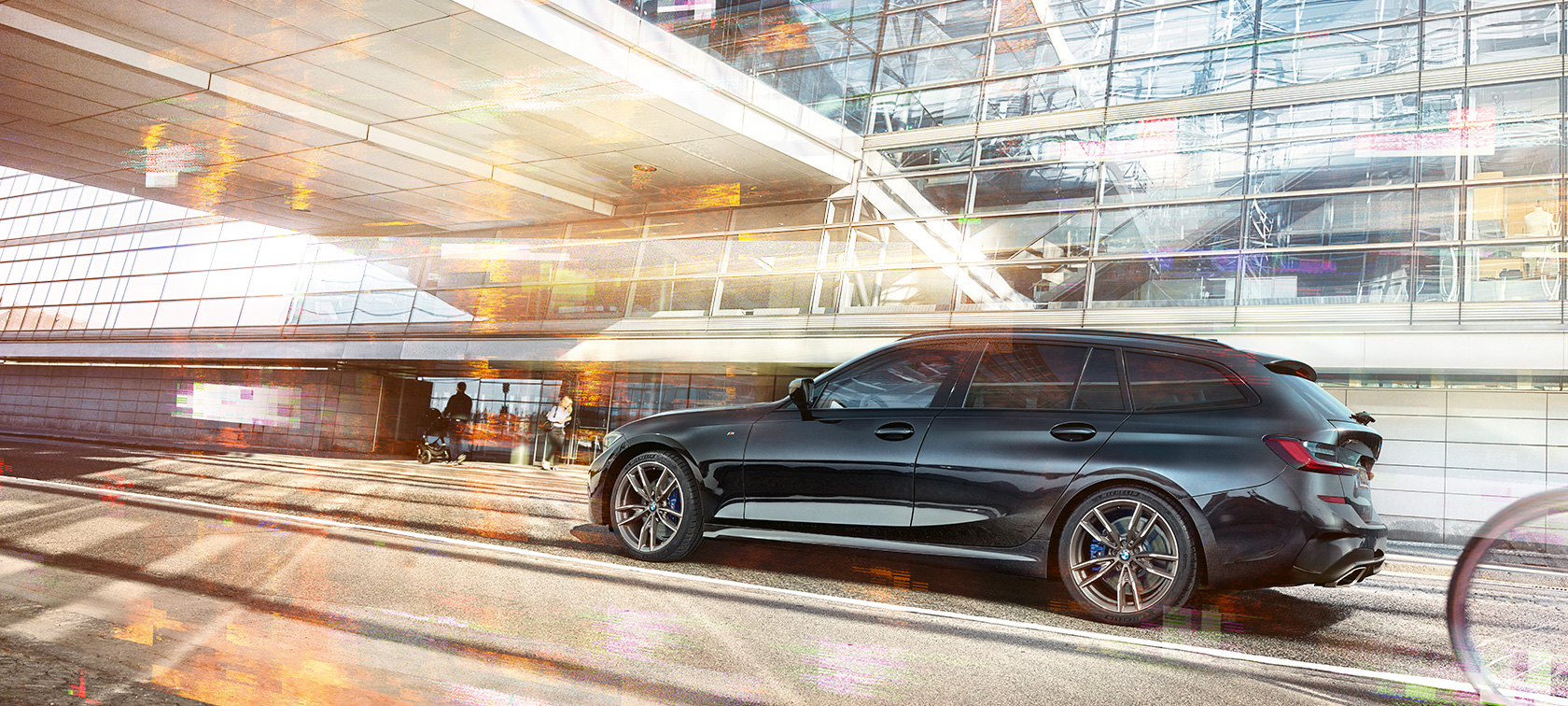 BMW M340i xDrive Touring mozgás közben, rézsútos elölnézetből Egy mozgó BMW M340i xDrive Touring rézsútos elölnézetből, egy üvegfalú városi épület előtt
