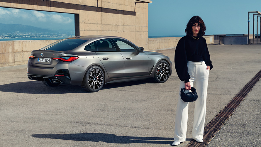 BMW M440i xDrive Gran Coupé, G26, 2021, egy modell társaságában látható autó rézsútos hátulnézetből BMW M440i xDrive Gran Coupé, G26, 2021, egy modell társaságában látható BMW Individual Frozen Pure Grey metálfényezésű autó rézsútos hátulnézetből