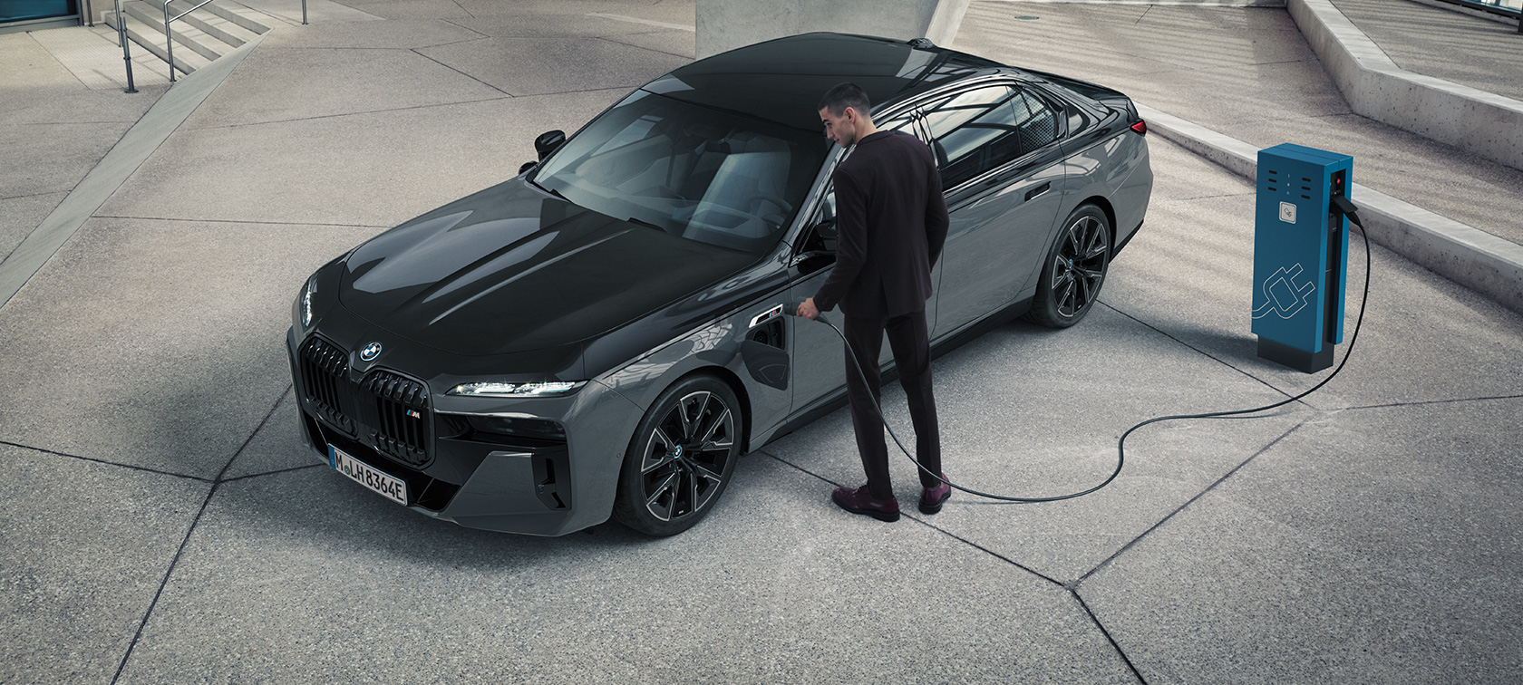 BMW M760e xDrive Limousine, G70, M Performance csomag BMW 760e xDrive Limousine, G70, M Performance csomag, BMW Individual kéttónusú Zafírfekete metál, BMW Individual Dravit szürke metál, egy töltőállomáson álló autó rézsútos elölnézetből egy töltőkábelt a kezében tartó férfival