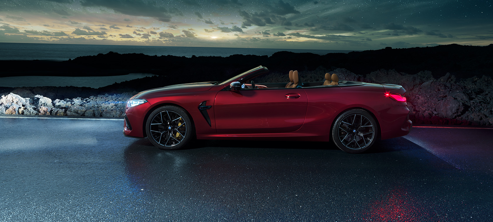 BMW M8 Competition Cabrio, oldalnézet. Egy parti úton, a csillagos ég alatt álló, nyitott vászontetős, Motegi vörös metálfényezésű BMW M8 Competition Cabrio oldalnézetből.