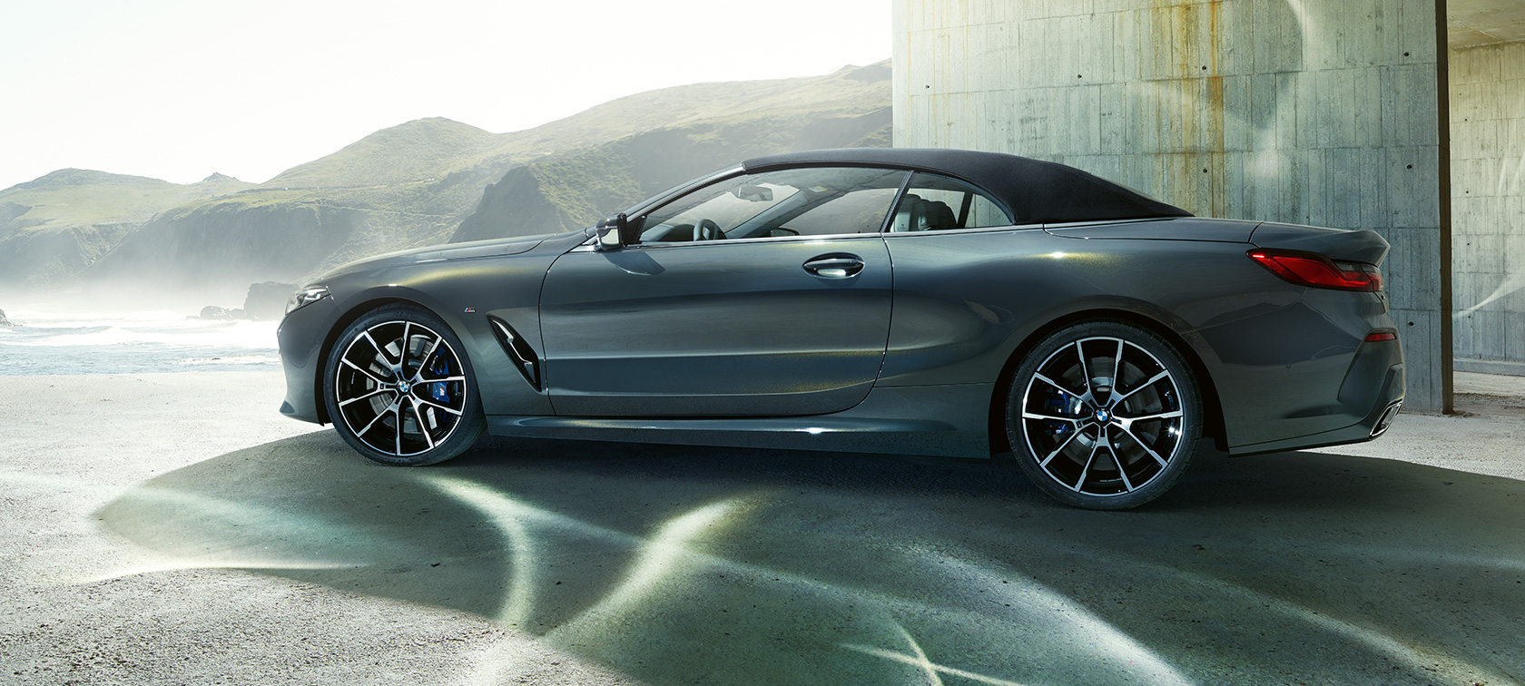 BMW M850i xDrive Cabrio, oldalnézet. Egy Dravit szürke metálfényezésű, nyitott vászontetős BMW M850i xDrive Cabrio hegyi tájjal a háttérben.