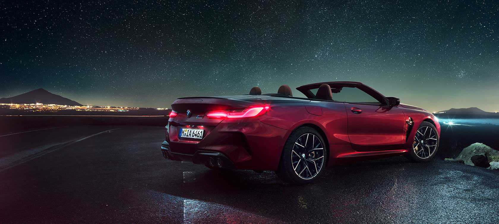BMW M8 Competition Cabrio, háromnegyedes hátulnézet. Egy, a csillagos ég alatt álló, nyitott vászontetős, Motegi vörös metálfényezésű BMW M8 Competition Cabrio háromnegyedes hátulnézetből.