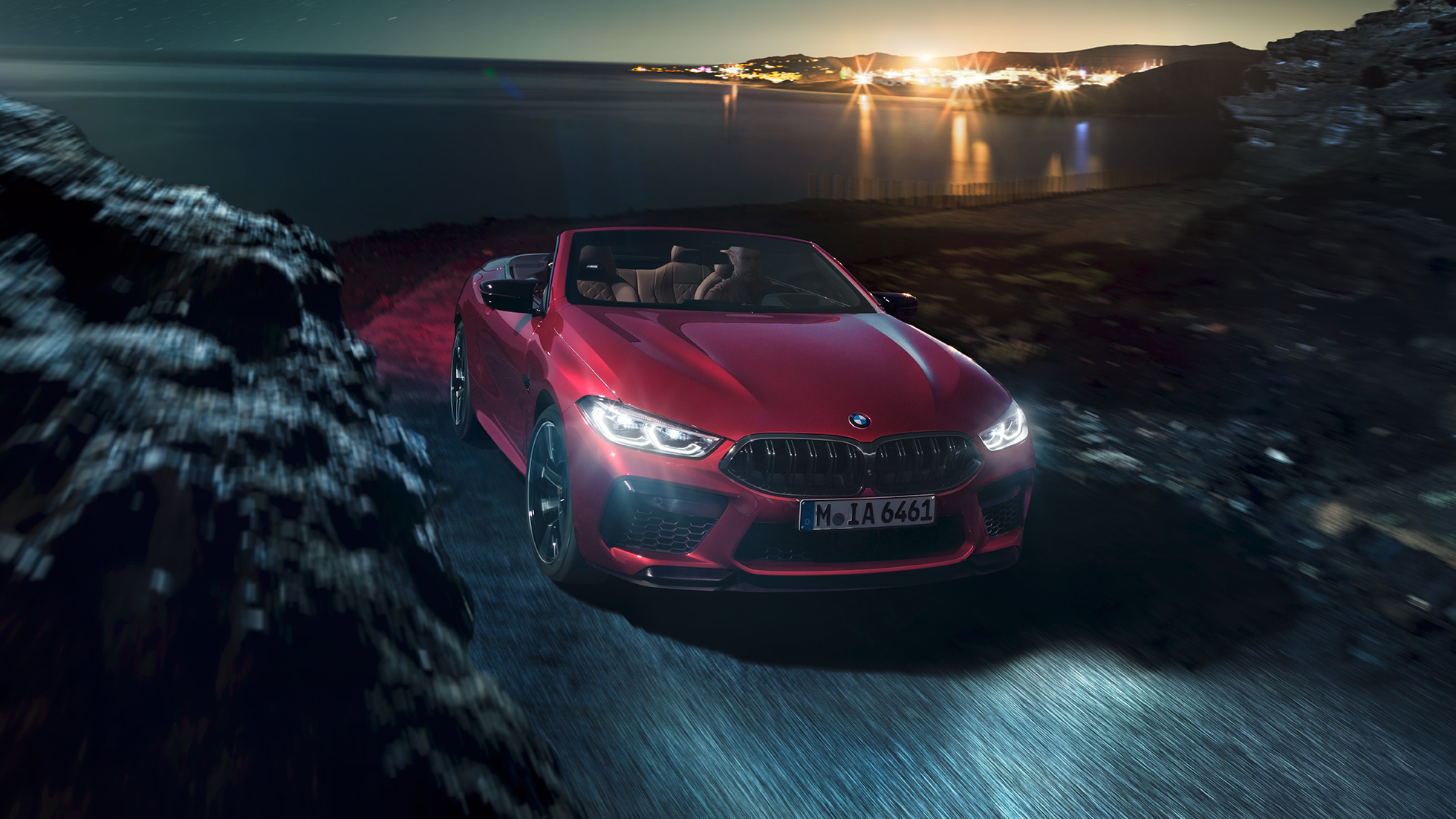 BMW M8 Competition Cabrio, háromnegyedes elölnézet. Egy éjszaka, a tengerparti úton nyitott vászontetővel suhanó, Motegi vörös metálfényezésű BMW M8 Cabrio háromnegyedes elölnézetből.