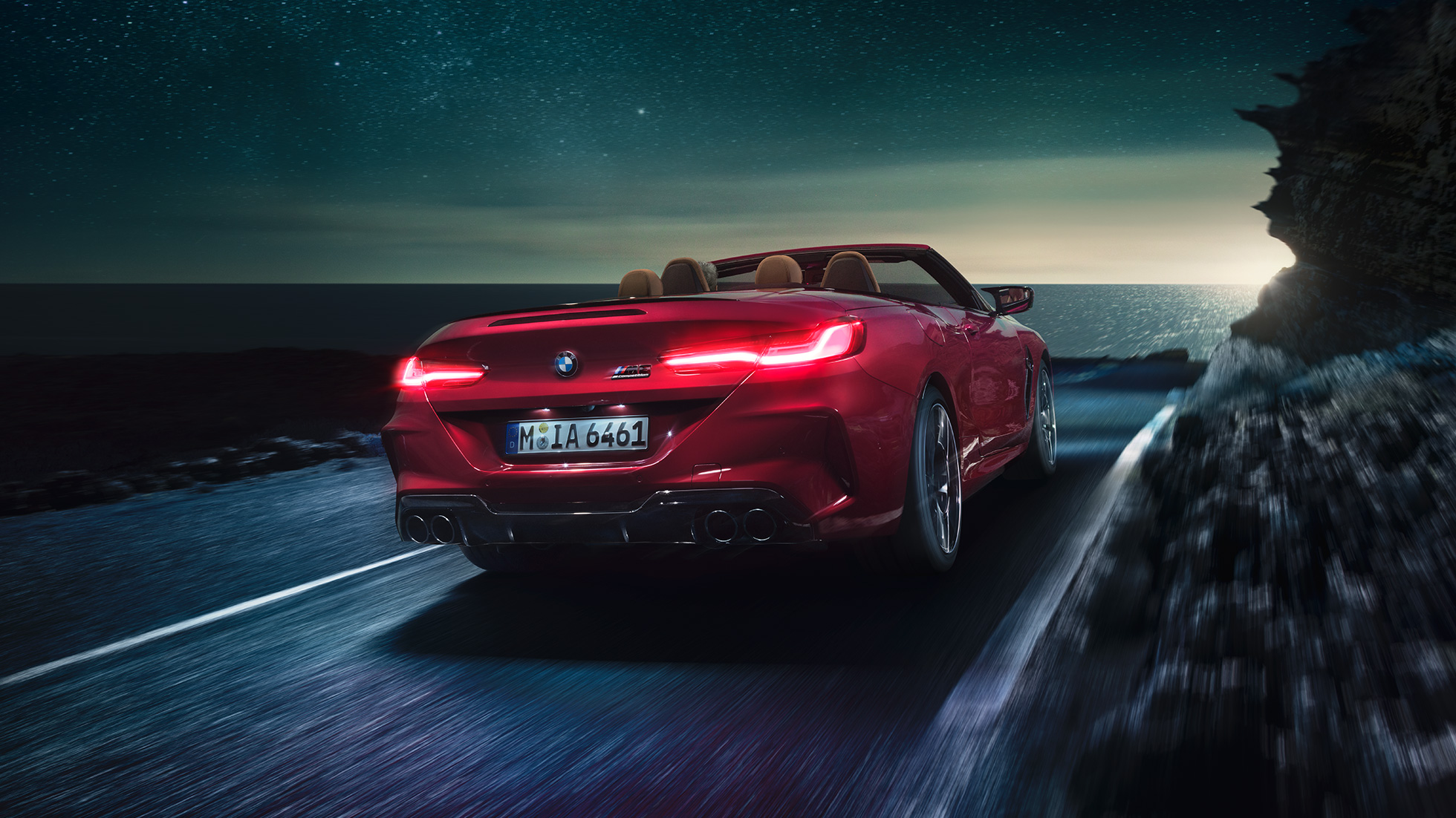 BMW M8 Competition Cabrio, háromnegyedes hátulnézet. Egy éjszaka, a tengerparti úton nyitott vászontetővel suhanó, Motegi vörös metálfényezésű BMW M8 Cabrio háromnegyedes hátulnézetből.