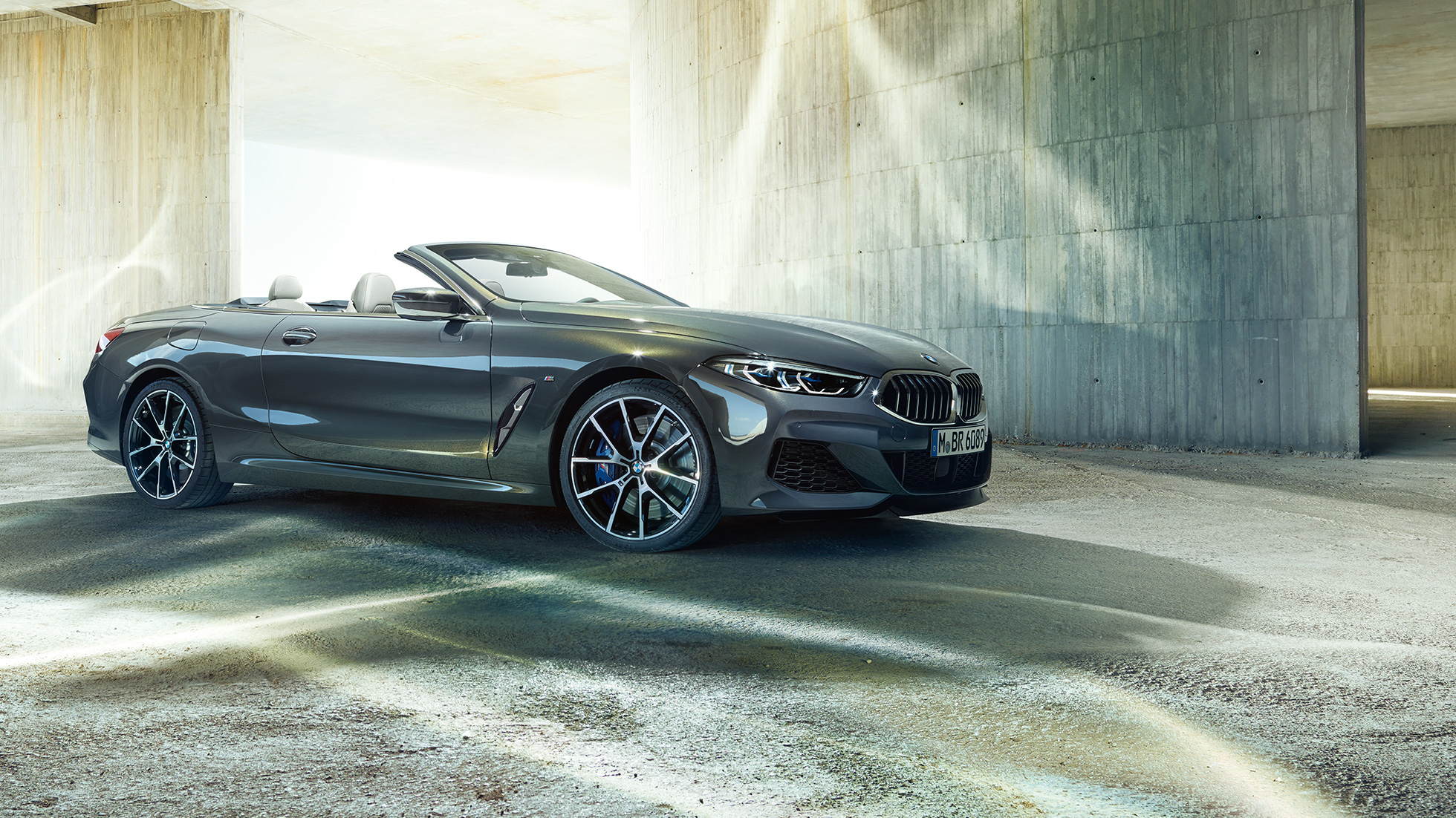 BMW M850i xDrive Cabrio, rézsútos oldalnézet. Egy betonépület előtt álló, nyitott vászontetős, Dravit szürke metálfényezésű BMW M850i xDrive Cabrio rézsútos oldalnézetből.