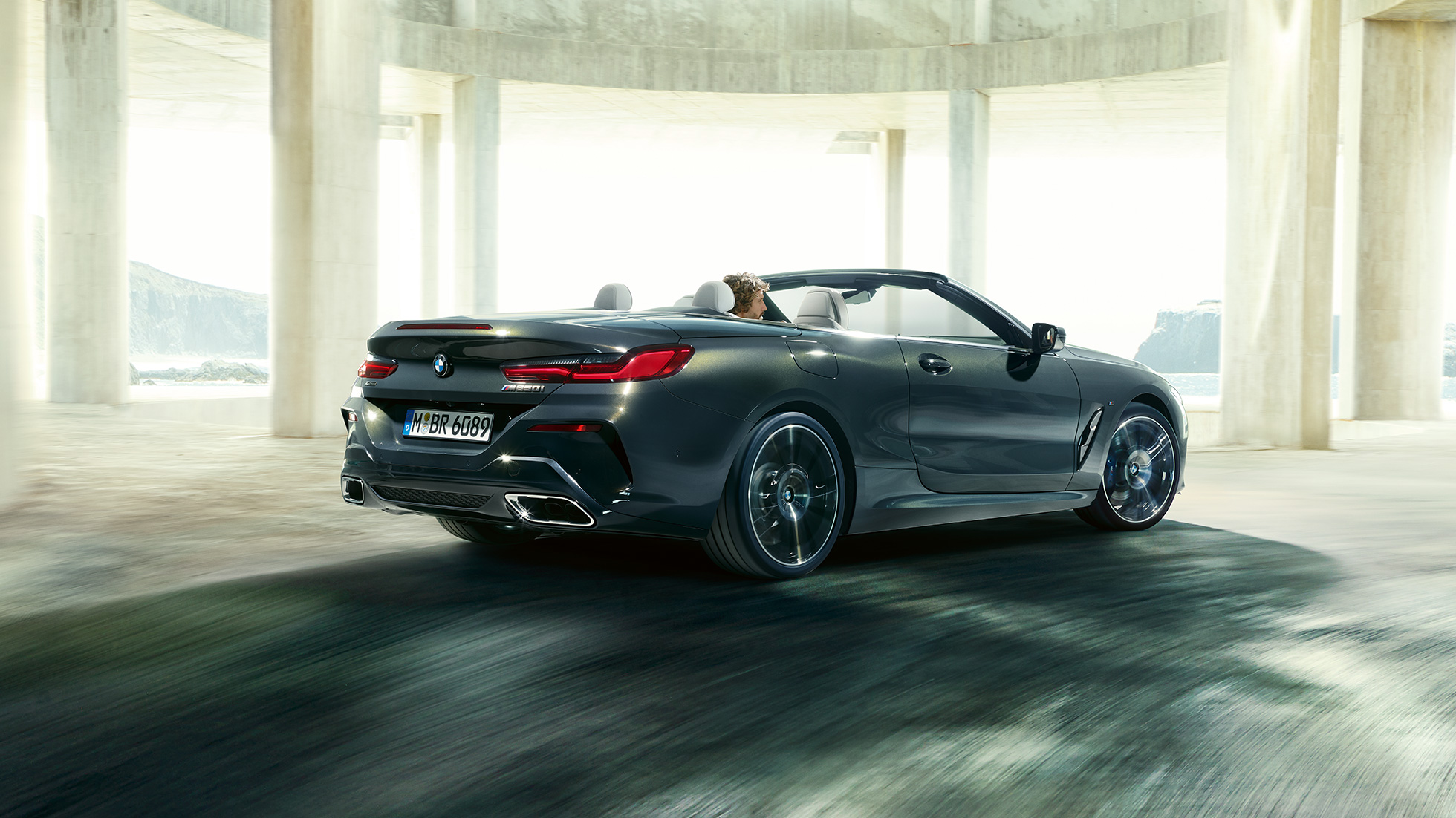 BMW M850i xDrive Cabrio, háromnegyedes hátulnézet. Egy betonépületen áthaladó, nyitott vászontetős, Dravit szürke metálfényezésű BMW M850i xDrive Cabrio háromnegyedes hátulnézetből.