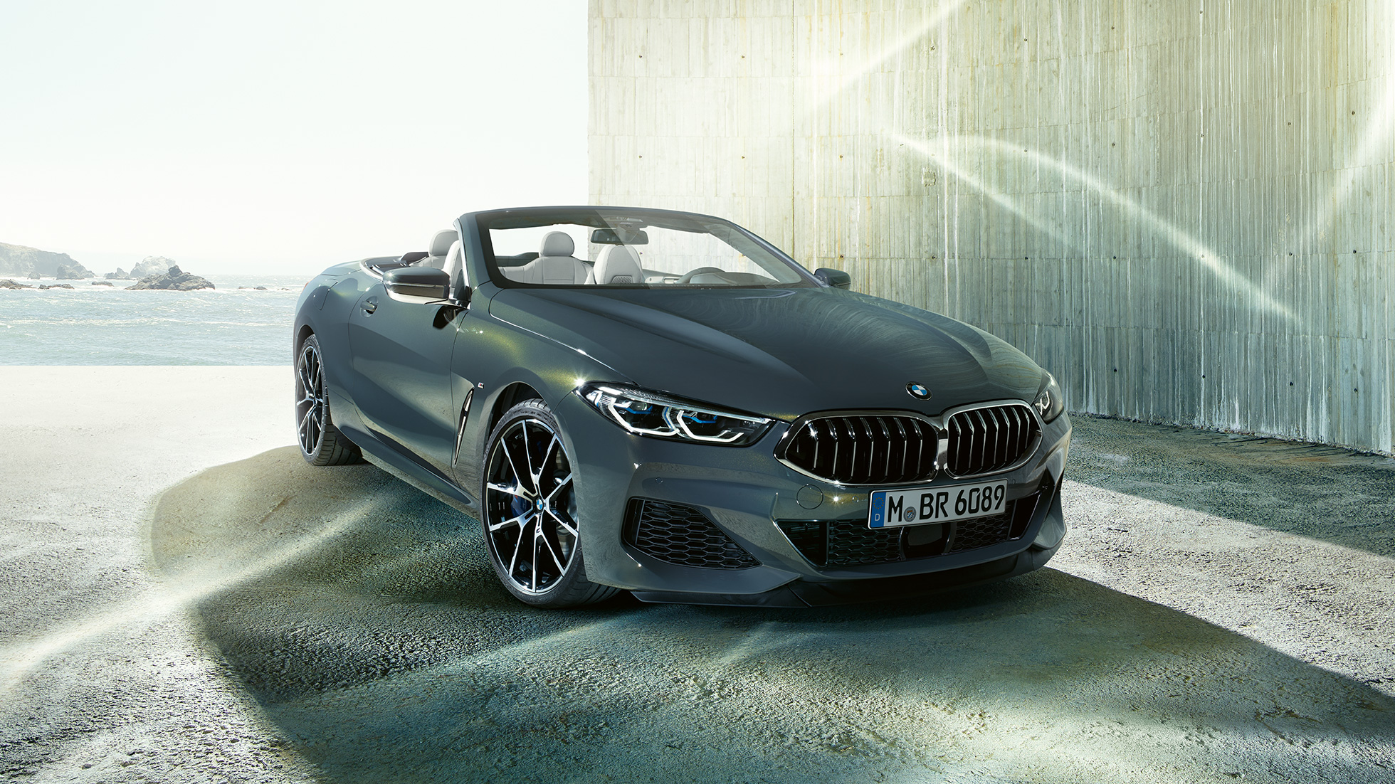 BMW M850i xDrive Cabrio, háromnegyedes elölnézet. Egy parton álló, nyitott vászontetős, Dravit szürke metálfényezésű BMW xDrive Cabrio háromnegyedes elölnézetből.
