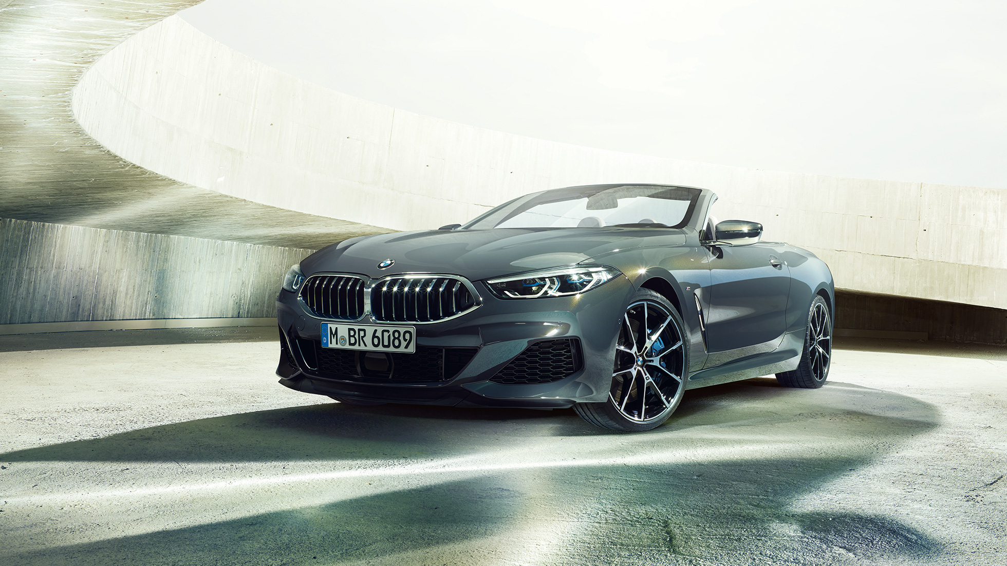 BMW M850i xDrive Cabrio, háromnegyedes elölnézet. Egy betonépület előtt álló, nyitott vászontetős, Dravit szürke metálfényezésű BMW M850i xDrive Cabrio háromnegyedes elölnézetből.