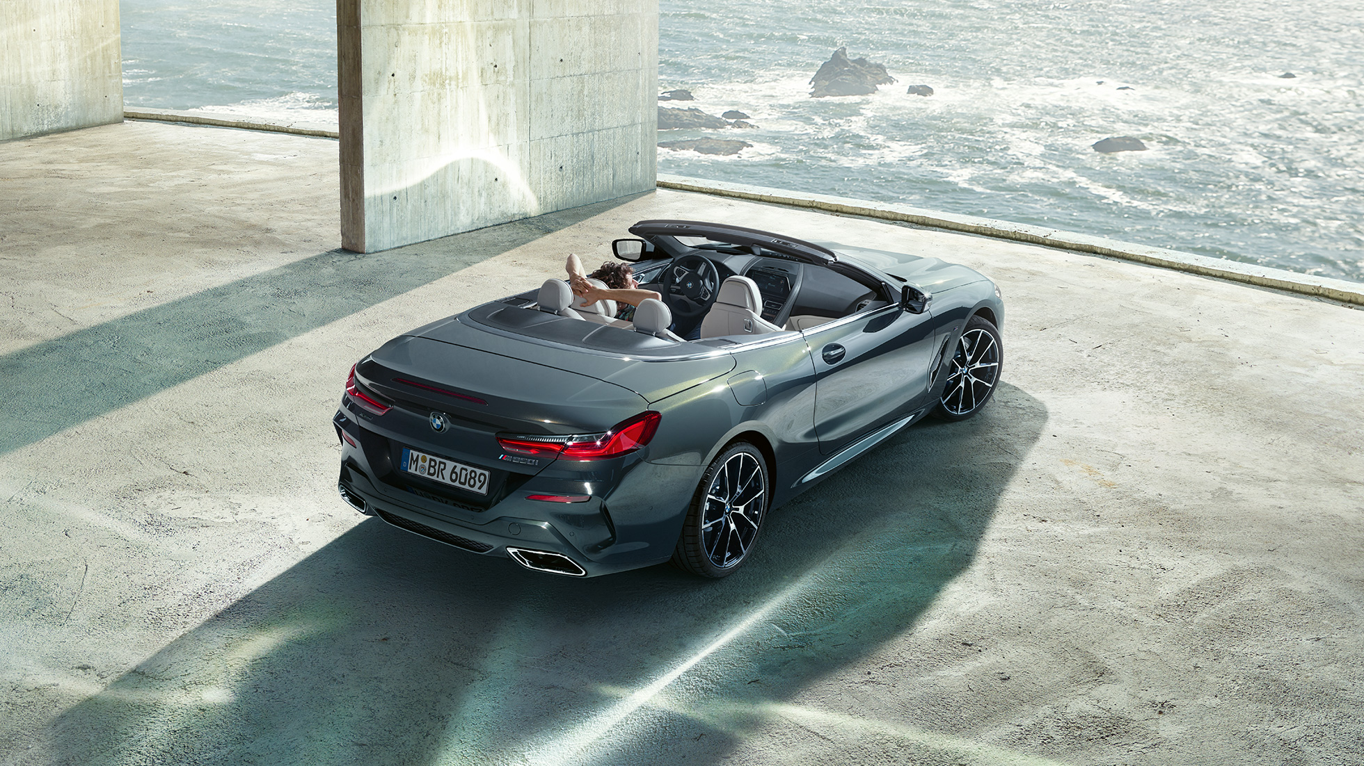 BMW M850i xDrive Cabrio, felülnézet. Egy óceánparton álló betonépületben parkoló, nyitott vászontetős, Dravit szürke BMW M850i xDrive Cabrio felülről.