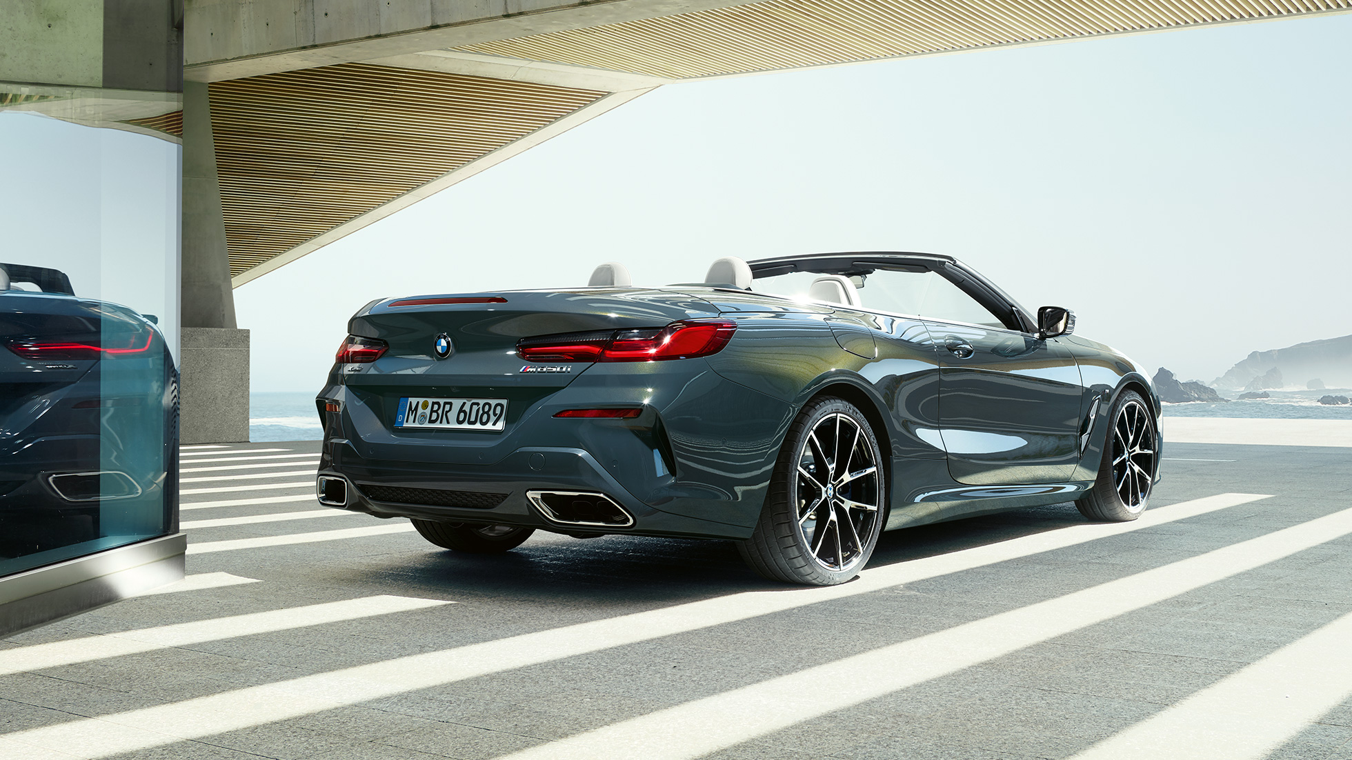 BMW M850i xDrive Cabrio, háromnegyedes hátulnézet. Egy álló, nyitott vászontetős, Dravit szürke metálfényezésű BMW M850i xDrive Cabrio háromnegyedes hátulnézetben, óceánnal a háttérben.