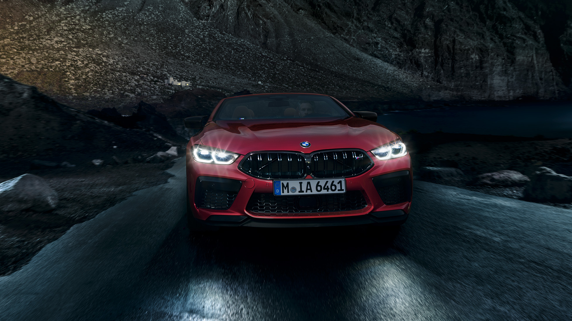 BMW M8 Competition Cabrio, elölnézet. Egy mozgó, világító fényszórójú, Motegi vörös metálfényezésű BMW M8 Competition Cabrio elölnézetből, éjszaka.
