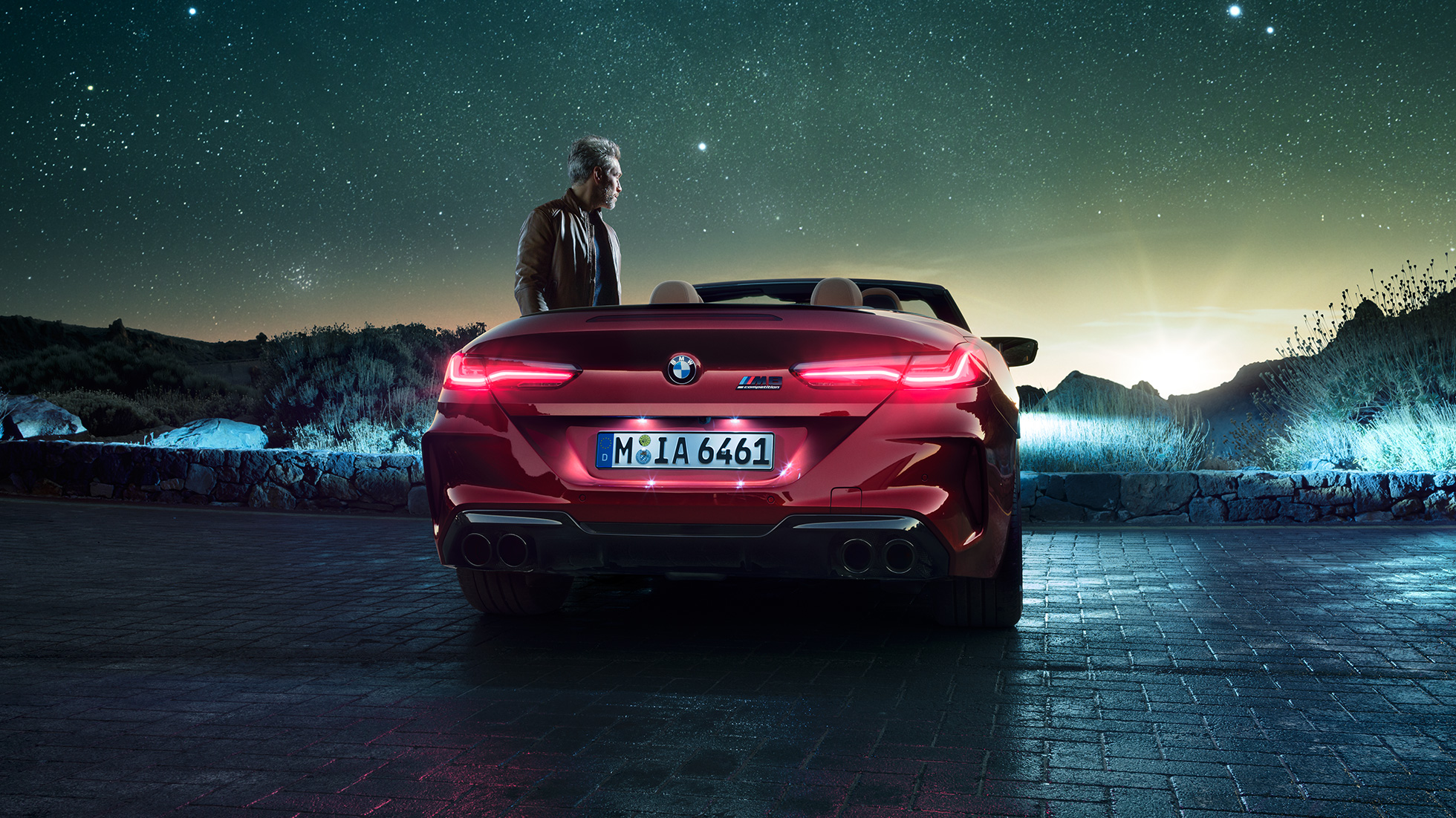 BMW M8 Competition Cabrio, hátulnézet. Egy nyitott vászontetős, Motegi Vörös BMW M8 Competition Cabrio hátulnézetből, egy csillagos ég alatt álló férfival.