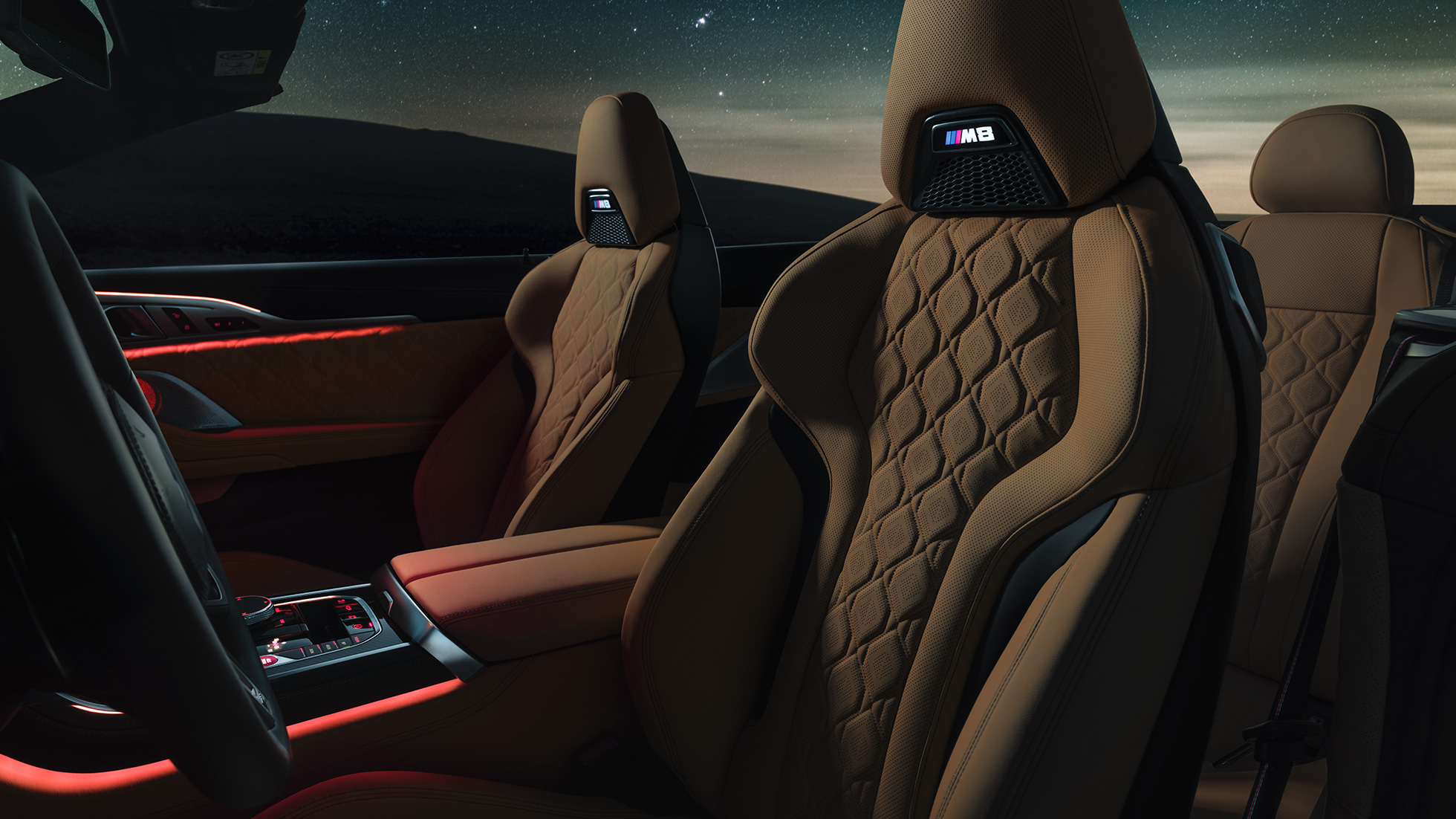 Merino bőrkárpitozású M Sport ülések. Egy, a csillagos ég alatt álló, nyitott tetős BMW M8 Competition Cabrio belső tere a Merino bőrkárpitozású M Sport ülésekkel és a vörös Hangulatvilágítással.