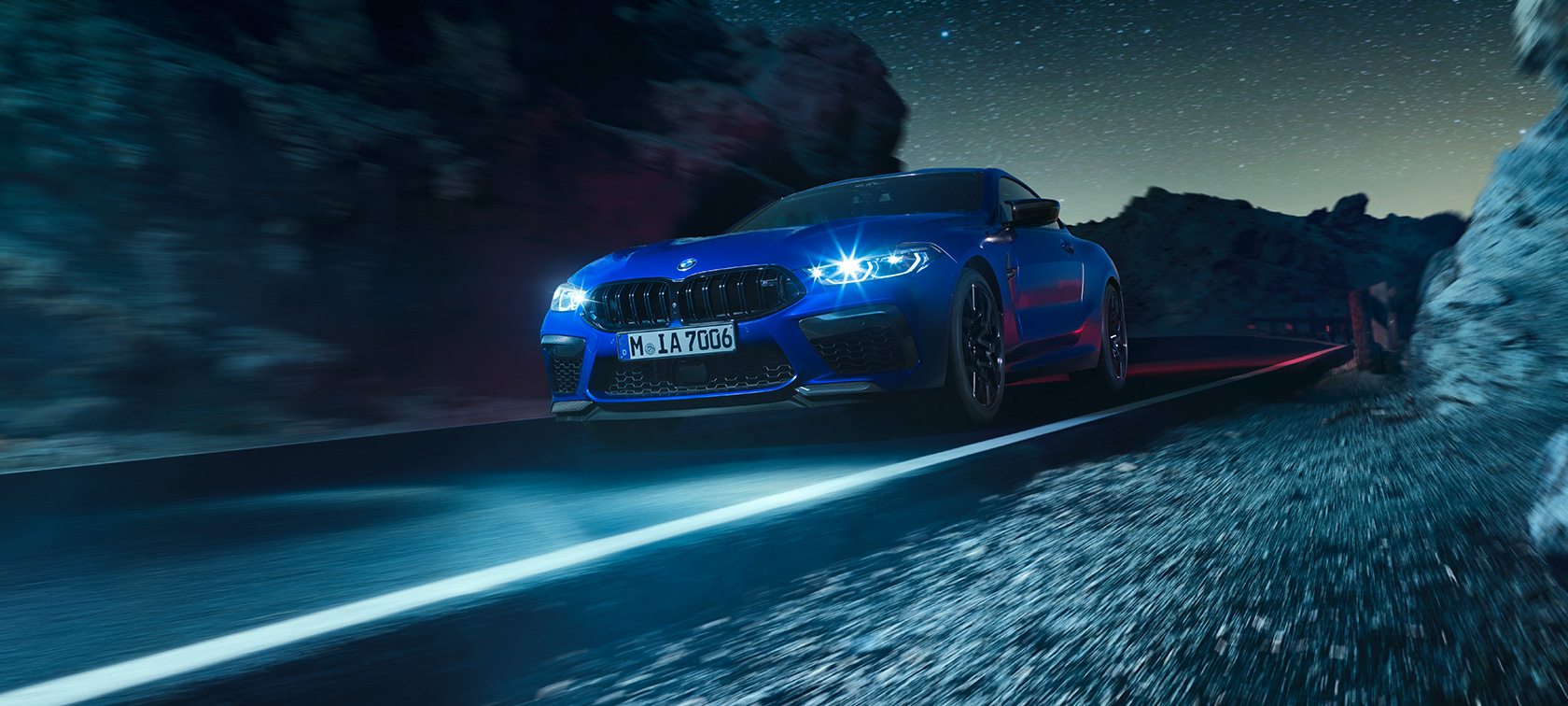 BMW M8 Competition Coupé, háromnegyedes elölnézet. Egy mozgó, Marina kék metálfényezésű BMW M8 Competition Coupé háromnegyedes elölnézetből, éjszaka.
