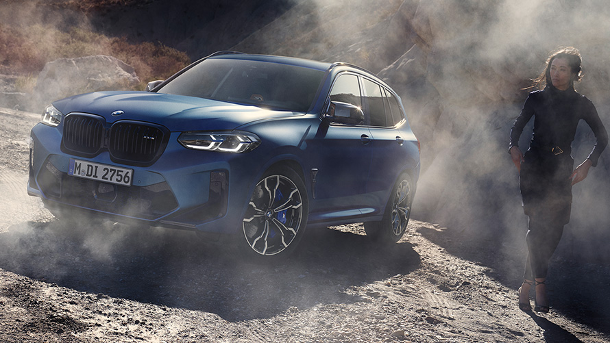 BMW X3 M Competition, F97 LCI, 2021, egy köves terepen álló autó rézsútos elölnézetből BMW X3 M Competition, F97 LCI, Facelift 2021, egy női modell társaságában látható, köves terepen álló Frozen Marina Bay kék metálfényezésű autó rézsútos elölnézetből