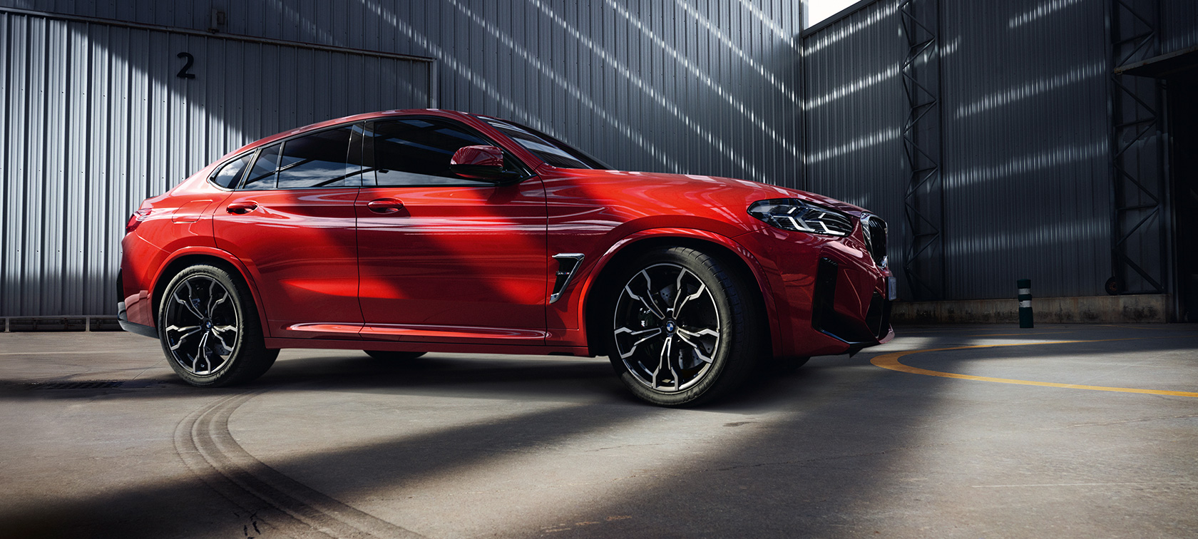 Rézsútos elölnézet, BMW X4 M, F98 LCI, Facelift 2021 BMW X4 M, F98 LCI, Facelift 2021, Toronto vörös metál, egy gyárépületben álló, rézsútos elölnézetből látható autó
