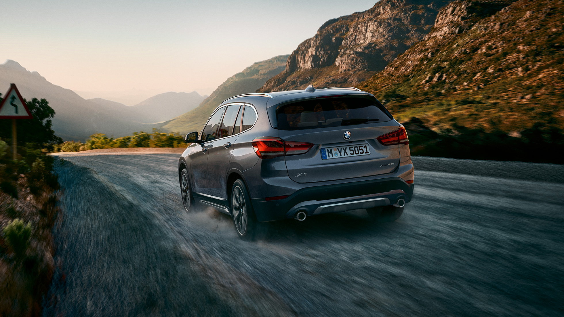 BMW X1, háromnegyedes hátulnézet. BMW X1, a mozgó autó háromnegyedes hátulnézetből, hegyvidéki tájjal a háttérben.