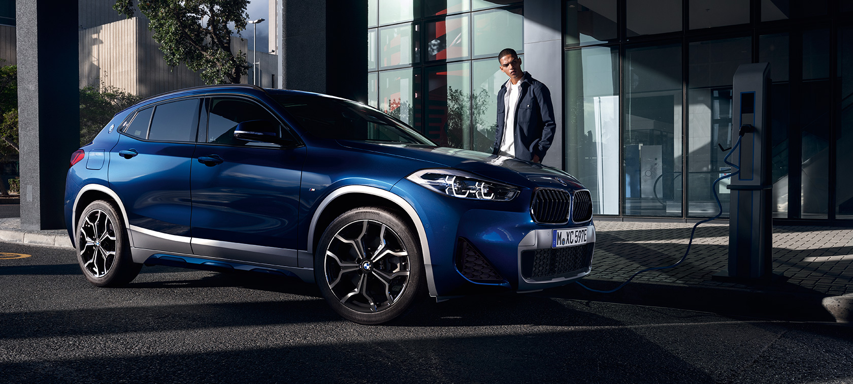 Plug-in hibrid, BMW X2 xDrive25e, F39, egy töltőállomáson tartózkodó autó rézsútos oldalnézetből Plug-in hibrid, BMW X2 xDrive25e, F39, egy töltőállomáson tartózkodó, modell társaságában látható autó rézsútos oldalnézetből