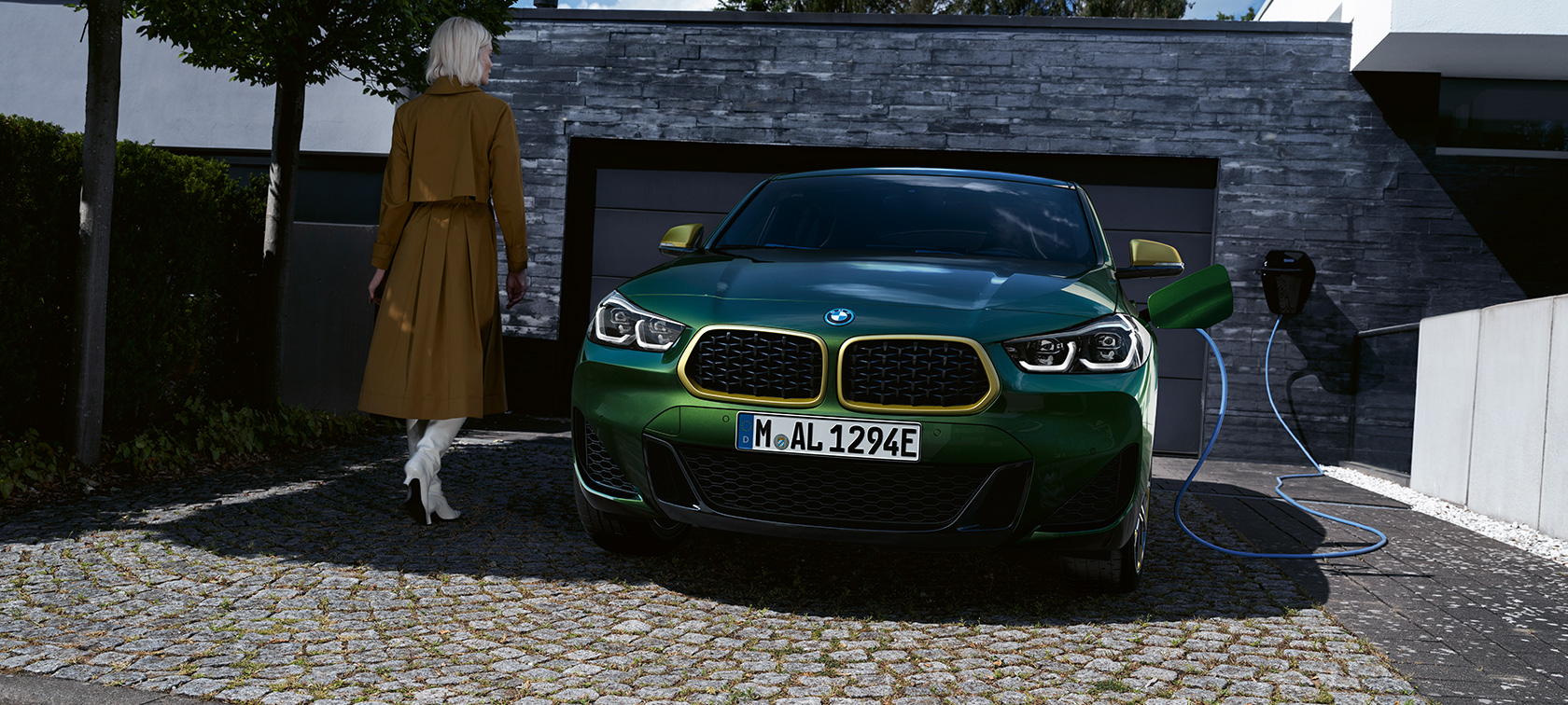 Elölnézet, BMW X2, F39, Sanremo zöld metál Egy töltés alatt álló autó, a bal oldalánál egy modellel, BMW X2, F39, Sanremo zöld metál