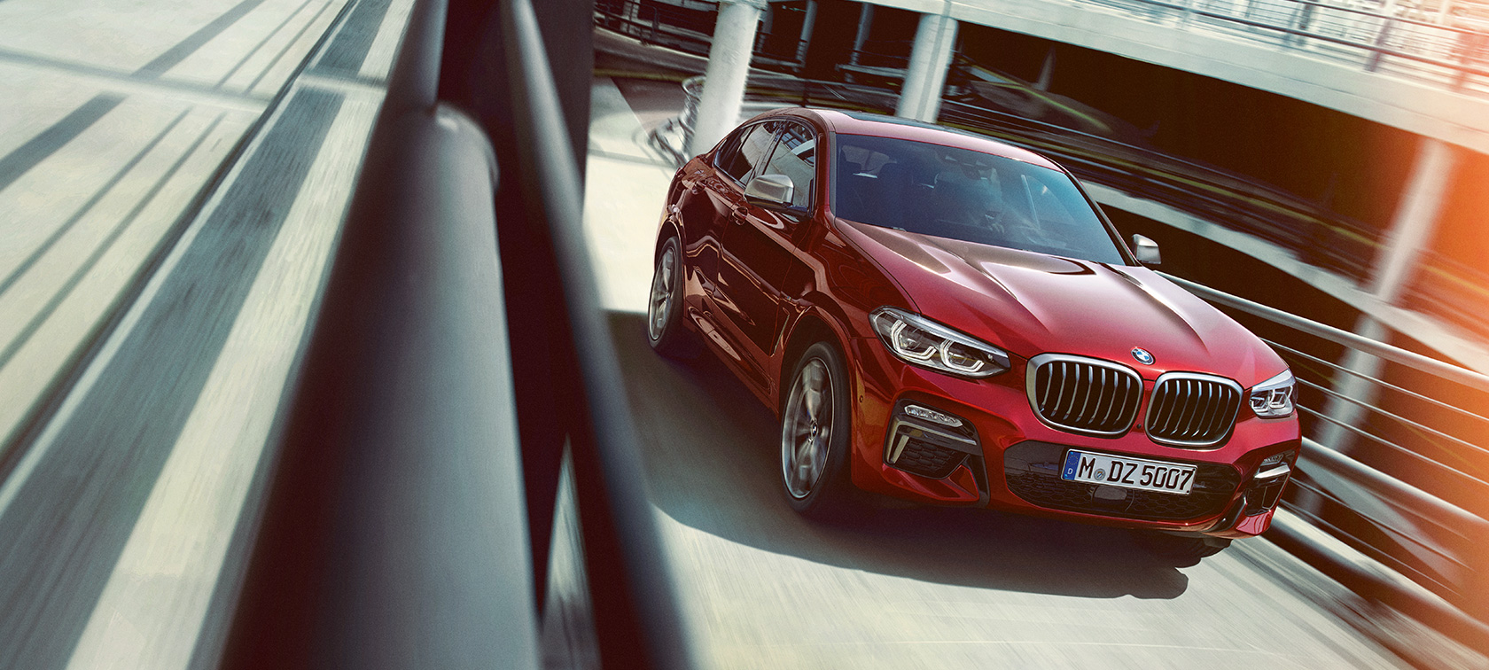 Egy többszintes parkolóházban haladó BMW X4 BMW X4 M40i, G02, 2018, Flamenco vörös briliáns hatással, egy többszintes parkolóházban haladó autó rézsútos oldalnézetből
