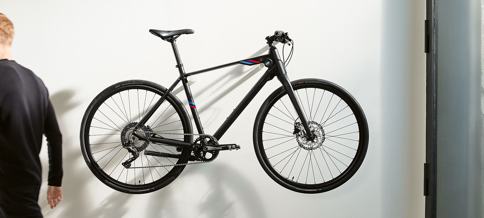 BMW M Bike, matt fekete. A képen egy, a fali tartókonzolra függesztett, a BMW M Motorsport csíkjait magán viselő, matt fekete BMW M Bike látható.