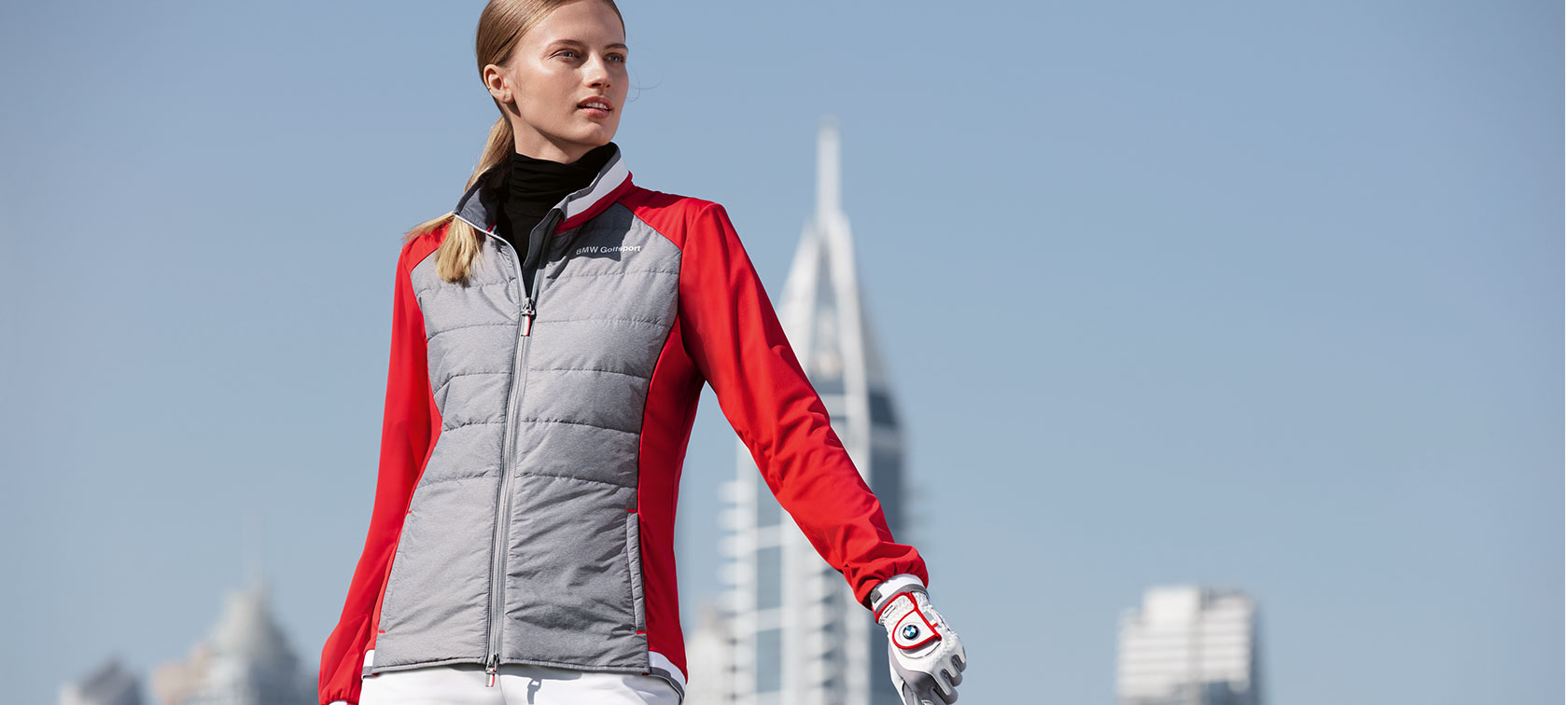 BMW Golfsport női dzseki. A képen egy, a BMW Golfsport Kollekció prémium darabjait bemutató fiatal nő látható, a háttérben egy nagyváros sziluettje.